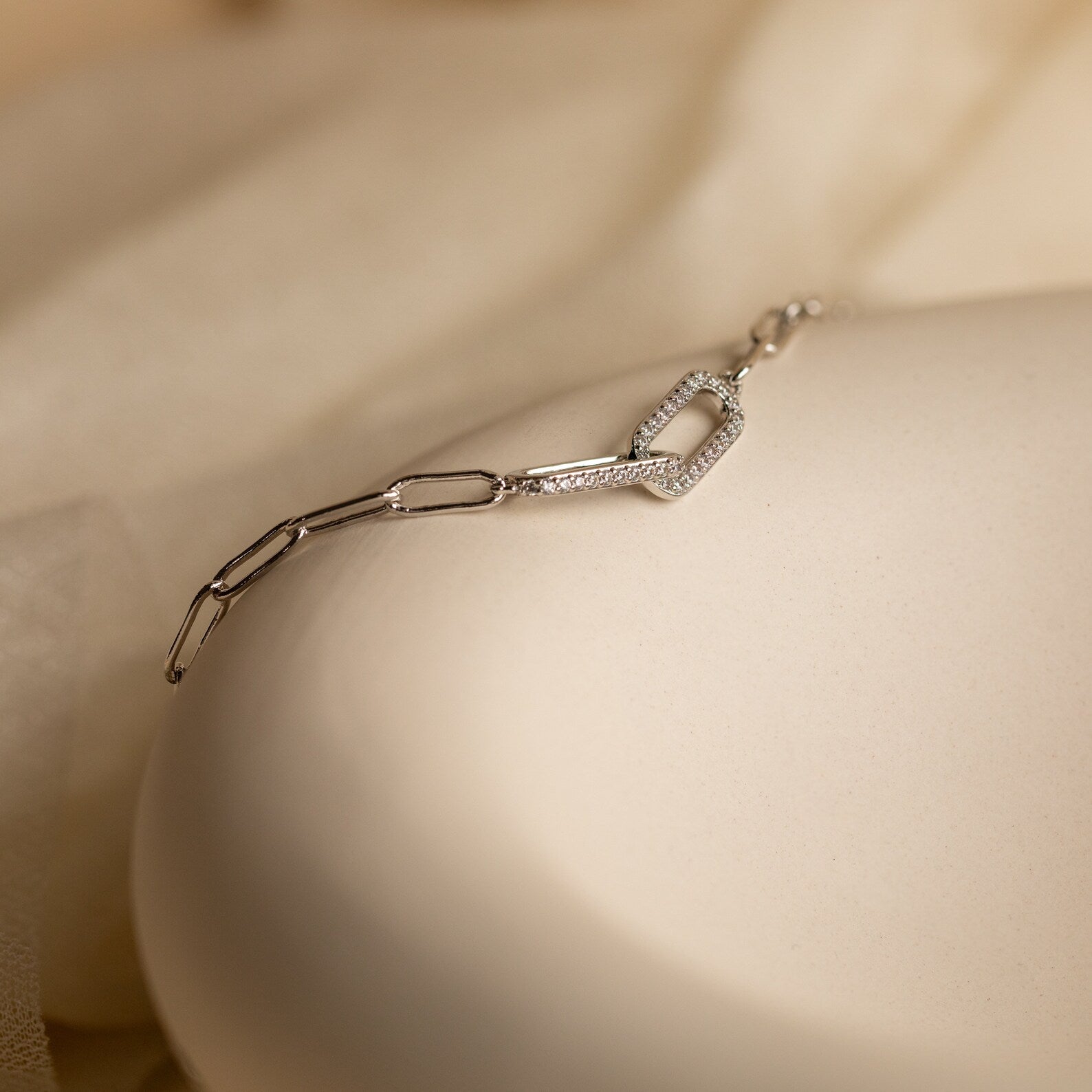 The Pave Link Chain Bracelet features delicate geometric links in silver, resting on a cream-colored surface—a minimalist and understated statement piece.