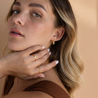A woman with wavy blonde hair wears Bold Ribbed Hoops and rings, touching her face while dressed in a brown top.