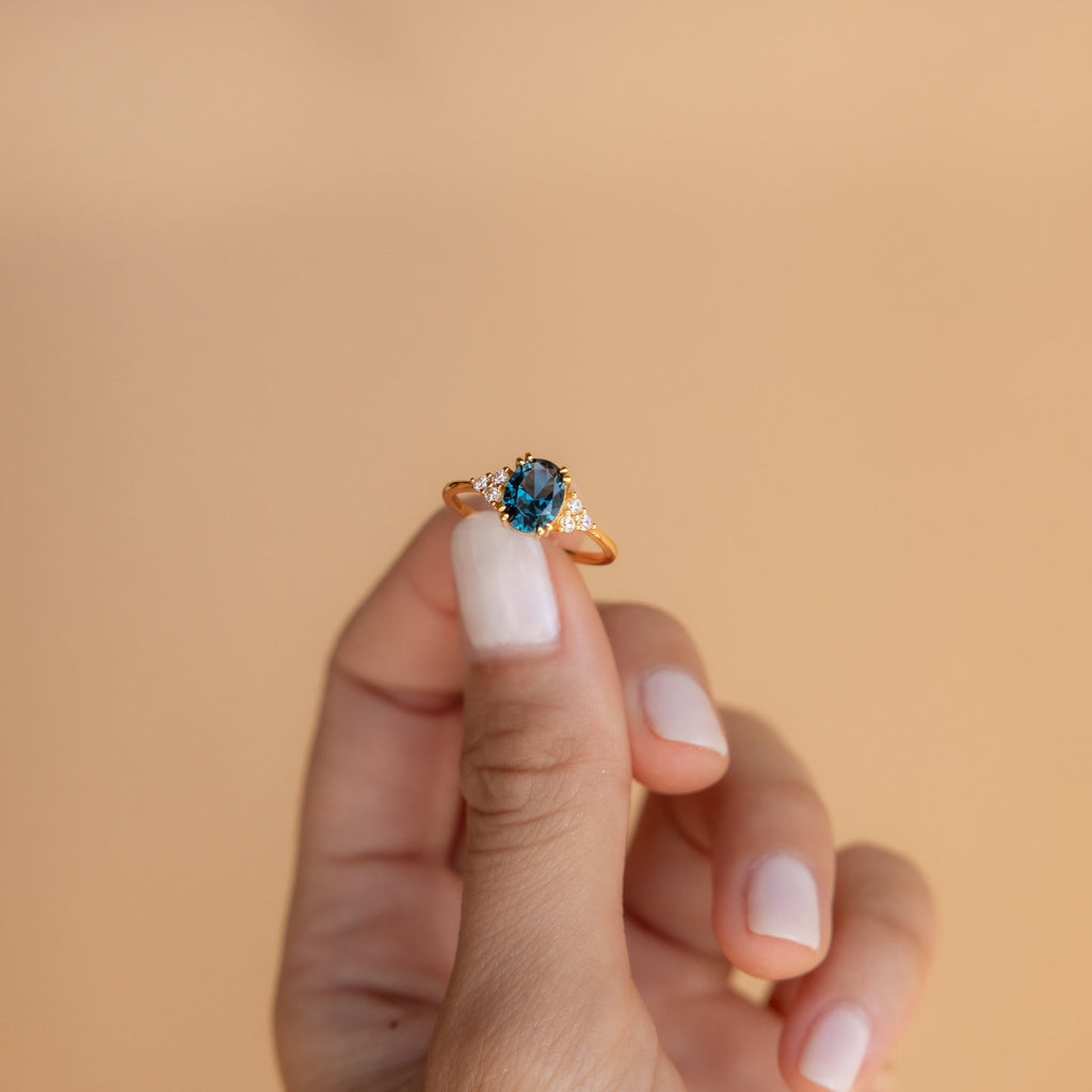 Hand holding a gold ring with a deep blue topaz center stone and side diamonds against a neutral background.