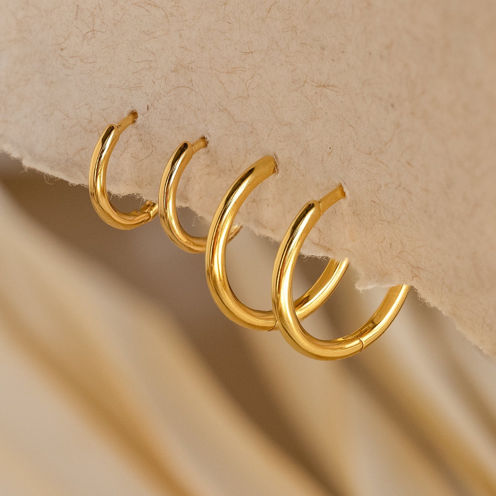 The Medium Bold Hoops in 18K Gold, designed for everyday wear, are showcased on a textured beige surface.