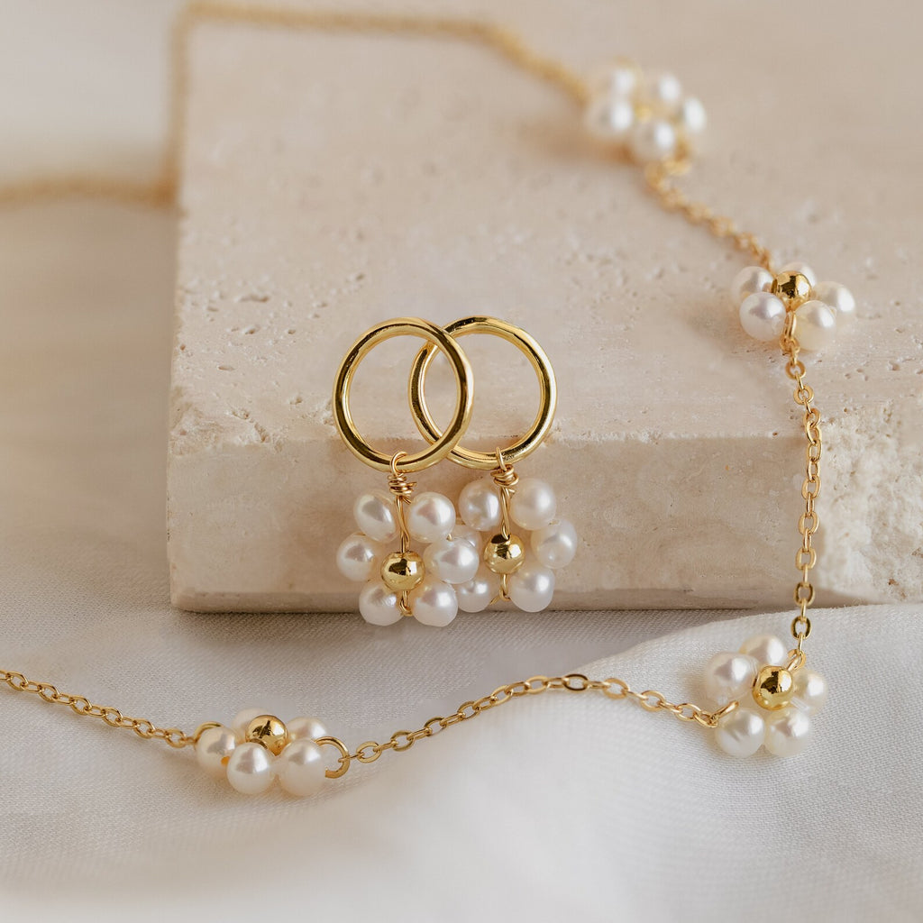 The Flower Pearl Station Necklace, displayed with gold hoop earrings on a beige stone slab, makes an elegant bridesmaids or Mother’s Day gift.