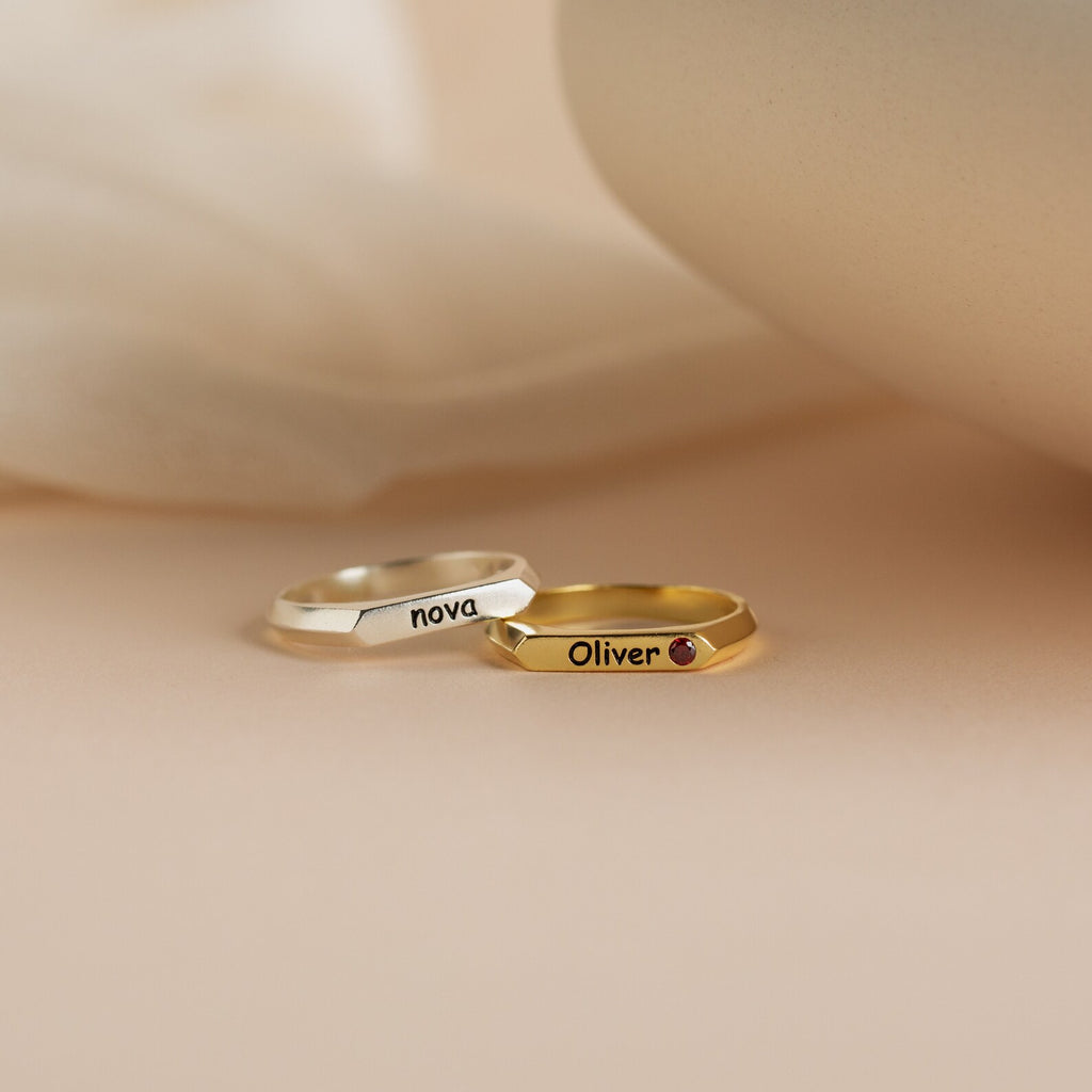 A Thin Name Signet Ring in silver with "Nova" and a gold ring featuring "Oliver" and a small gem are displayed on a soft beige surface.
