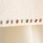 Nine pairs of Duo Birthstone Studs, featuring small colorful stones, are lined up on a cream fabric background—an ideal bridesmaids gift or a personalized touch for your everyday style.