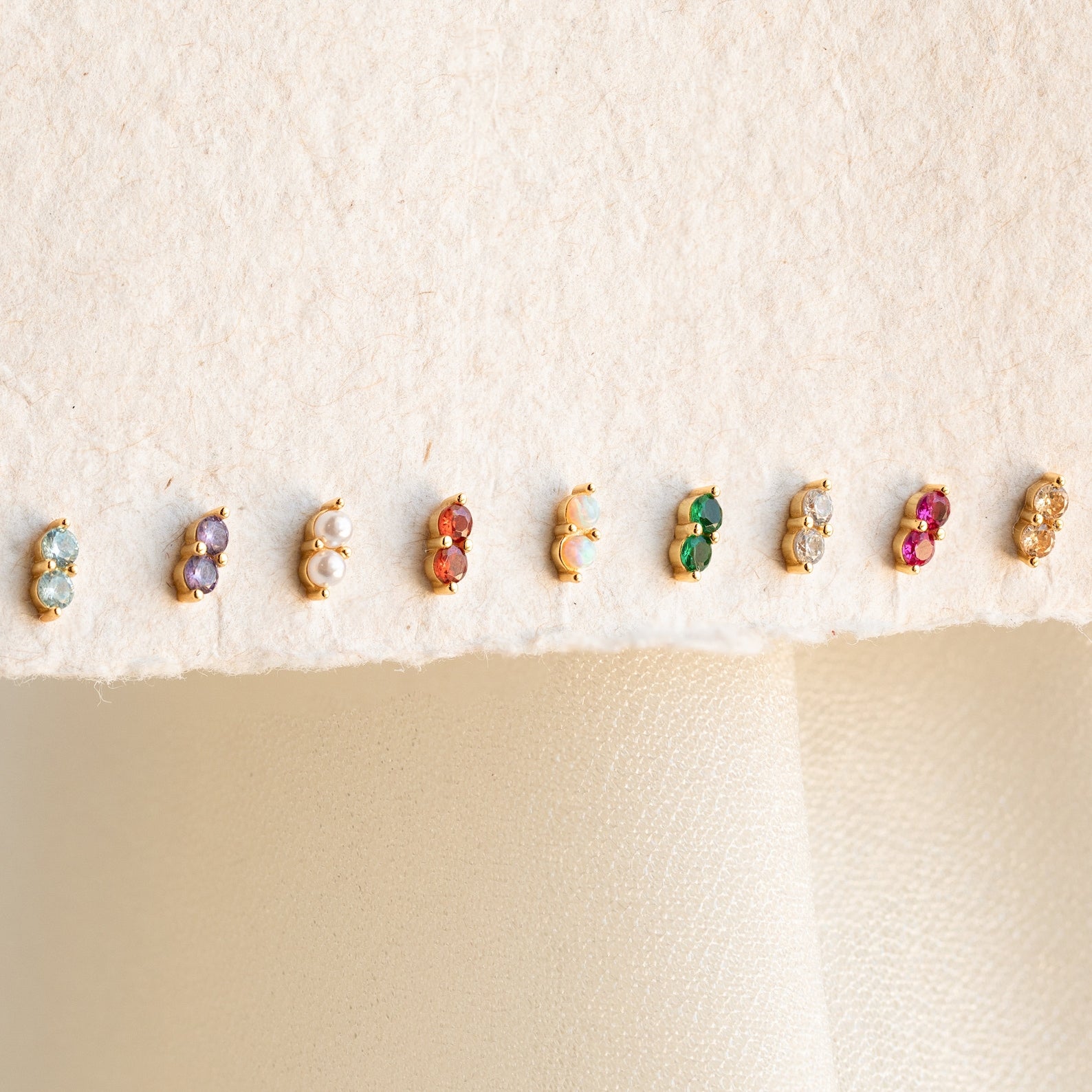 Nine pairs of Duo Birthstone Studs, featuring small colorful stones, are lined up on a cream fabric background—an ideal bridesmaids gift or a personalized touch for your everyday style.