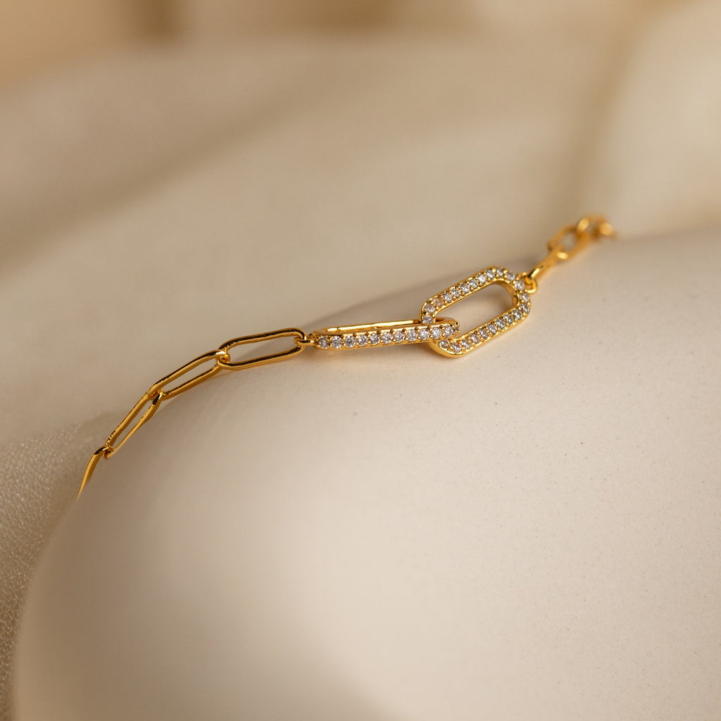 The Pave Link Chain Bracelet features a gold chain with a diamond-studded link, shown on a soft beige surface—a striking addition to any jewelry collection.