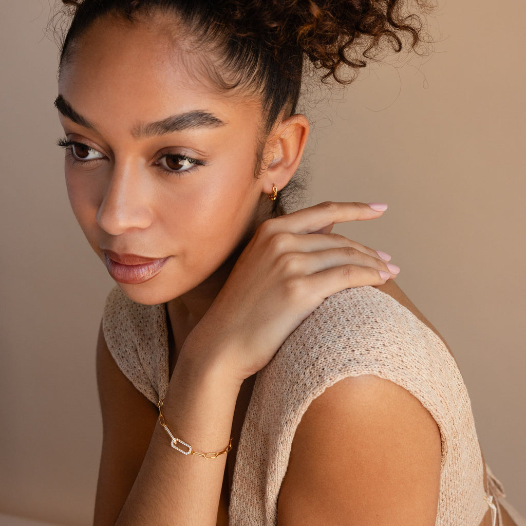 A woman with curly hair wears a sleeveless knit top and gold jewelry, including the Pave Link Chain Bracelet, as she looks thoughtfully to the side.