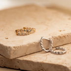 Two pairs of Inside Out Diamond Huggies, elegant hoop earrings in gold and silver with sparkling stones, displayed on beige stone slabs.