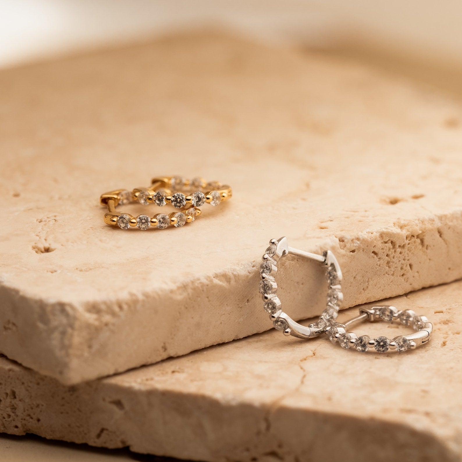 Two pairs of Inside Out Diamond Huggies, elegant hoop earrings in gold and silver with sparkling stones, displayed on beige stone slabs.