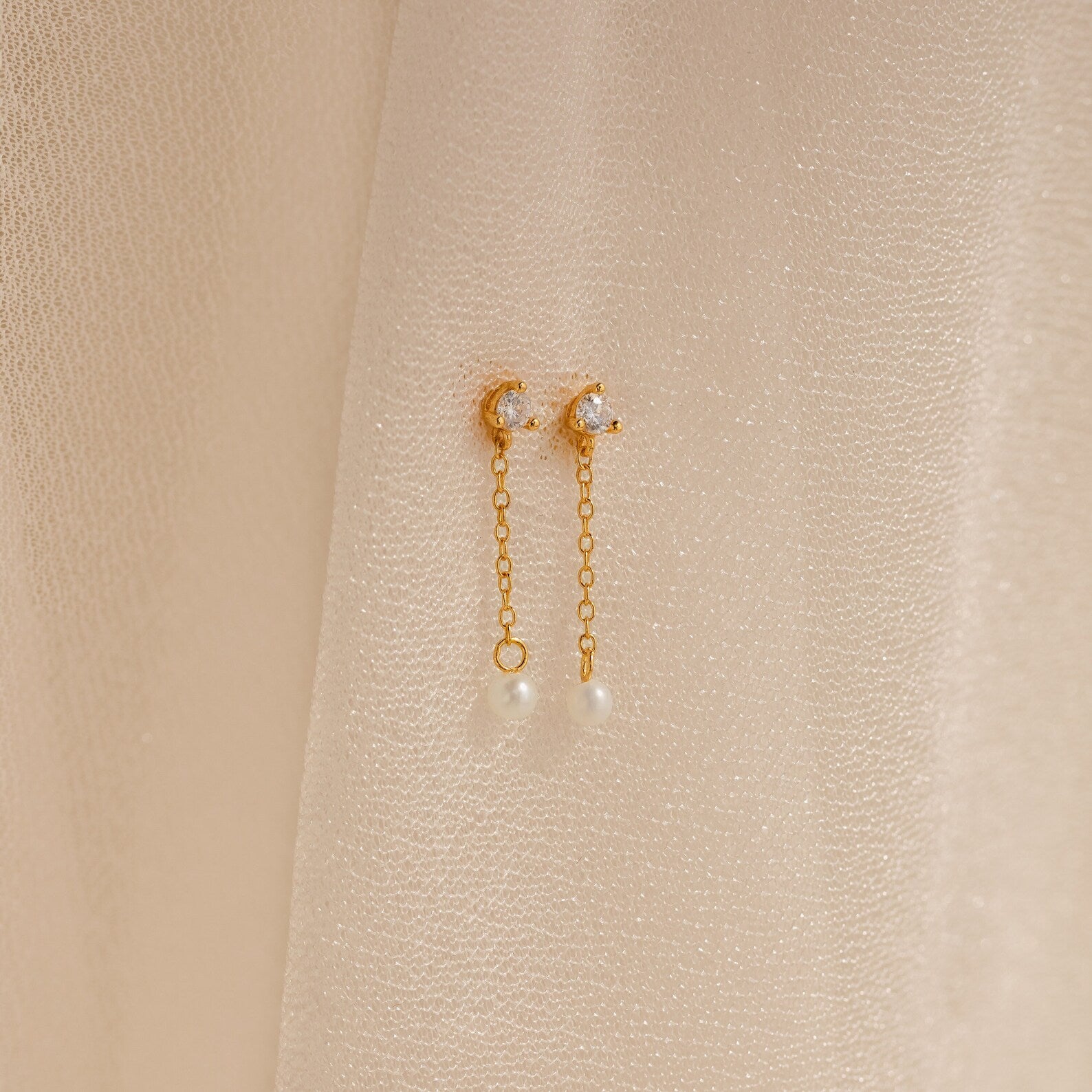 Diamond Pearl Drop Earrings with crescent moons, chain drops, and pearls are displayed on a soft beige fabric background, highlighting their timeless elegance.