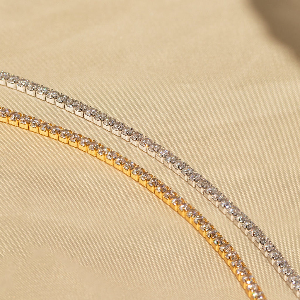 The Classic Diamond Tennis Bracelet, available in gold and silver, features sparkling gemstones and a timeless design—an elegant jewelry piece displayed on a beige fabric background.