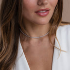 Woman with wavy brown hair wears a white V-neck top and the Dainty Diamond Tennis Necklace & Bracelet Set in White Gold, featuring a thin, sparkling choker reminiscent of a classic diamond tennis style.