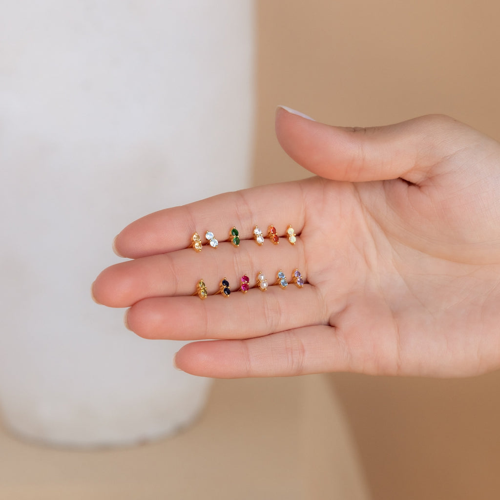 A hand displays nine Duo Birthstone Studs in vibrant colors, arranged in three neat rows—ideal as a thoughtful bridesmaids gift or to add a personalized gemstone accent to any jewelry collection.