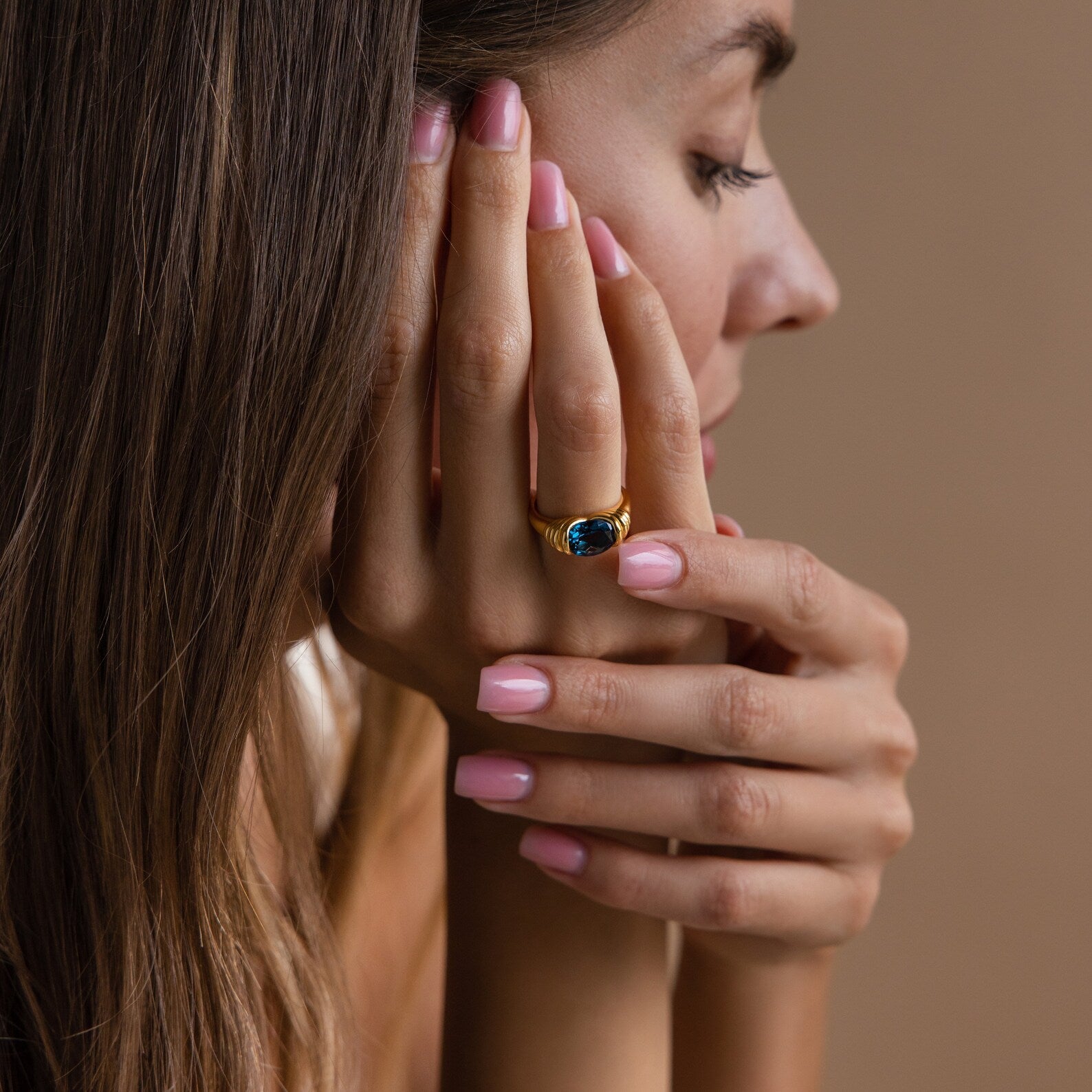 A woman with pink nails, wearing the London Blue Signet Ring with a gold band and London Blue Topaz stone, touches her face.