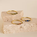 The Morse Code Birthstone Ring and two other gold gemstone rings are displayed on two stacked beige stone slabs.