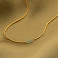 Close up of a dainty gold necklace featuring a small horizontal jade bar pendant, displayed on a soft fabric background.
