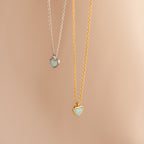 Two heart-shaped necklaces—one silver, one gold—with light blue stones rest on a beige background. Add the Tiny Opal Heart Necklace to your collection for a touch of charm.