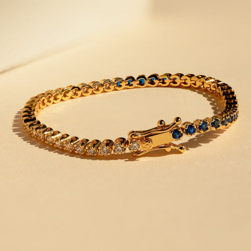 The Blue Diamond Tennis Bracelet features alternating clear and blue gemstones for an elegant, minimalist look—perfect as your "something blue." Shown on a beige surface under warm lighting.