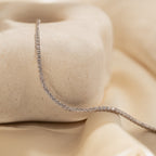 The Dainty Diamond Tennis Necklace & Bracelet Set in White Gold is elegantly displayed draped over a smooth stone and beige satin fabric.