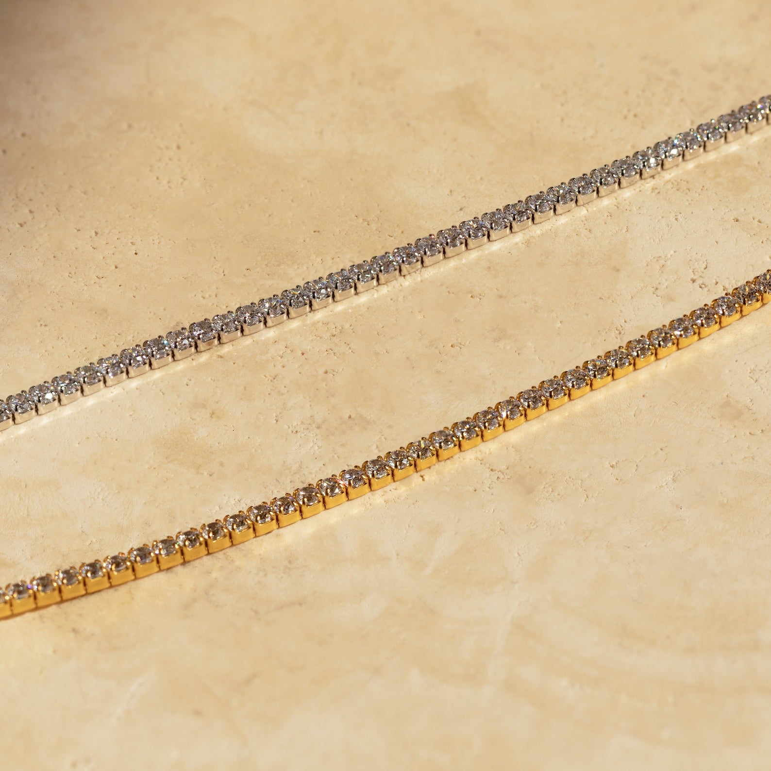 Close-up of two diamond tennis choker necklaces displayed on a sunlit stone surface—one with a white gold setting and the other with a yellow gold setting, both showcasing round-cut diamonds in a prong setting.