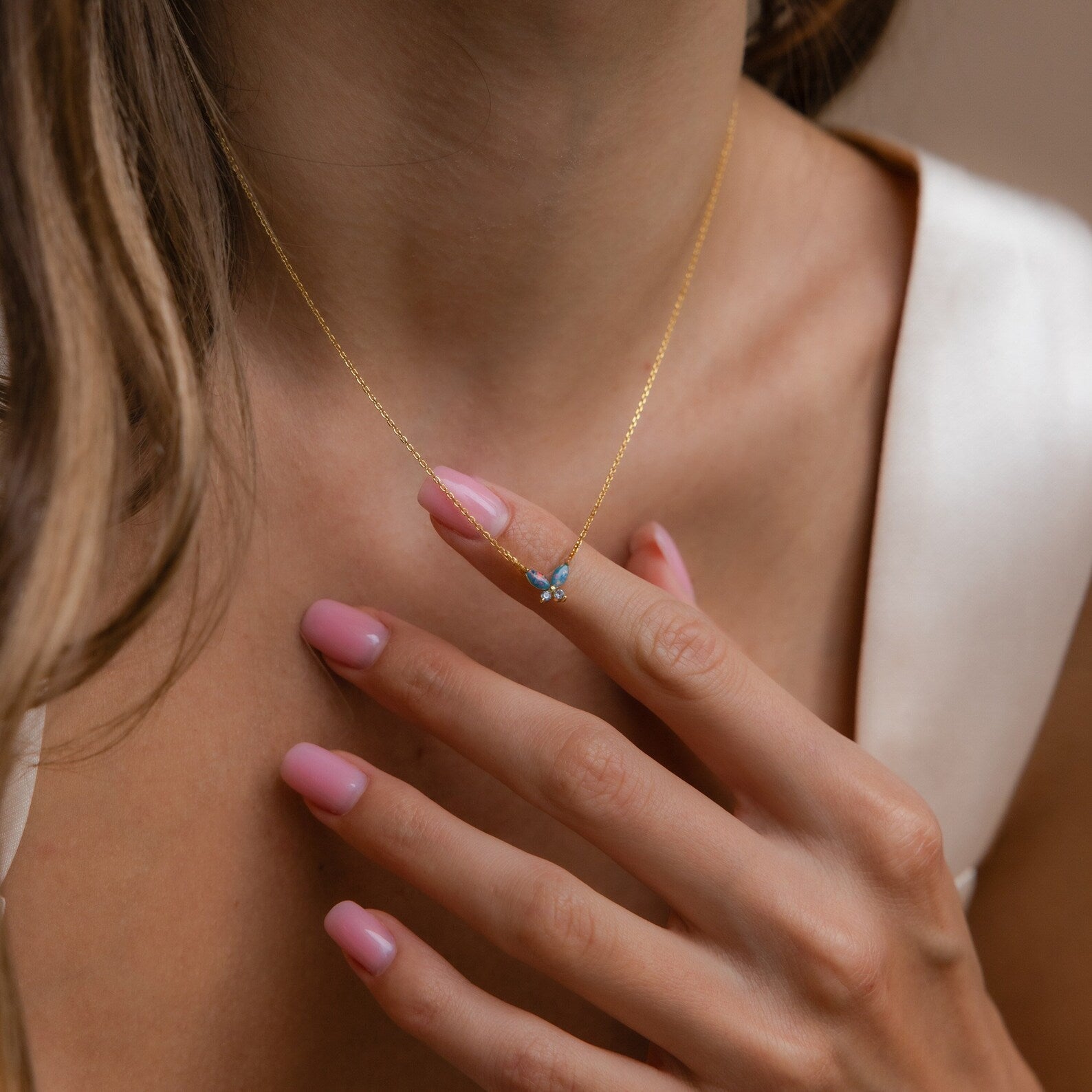 A woman with pink-manicured nails gently touches her Blue Opal Butterfly Necklace, showcasing its delicate butterfly pendant.