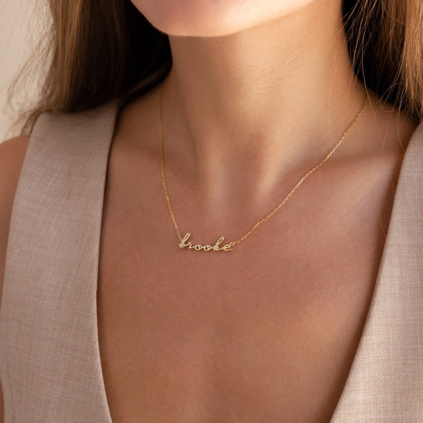 A woman wears the Dainty Bubble Name Necklace personalized with "Brooke" in gold cursive, styled with a beige sleeveless top.