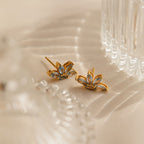 Aquamarine Marquise Studs with gold and blue floral design rest on cream fabric beside a clear glass object—ideal as a bridesmaids gift or for adding elegance to any special occasion.