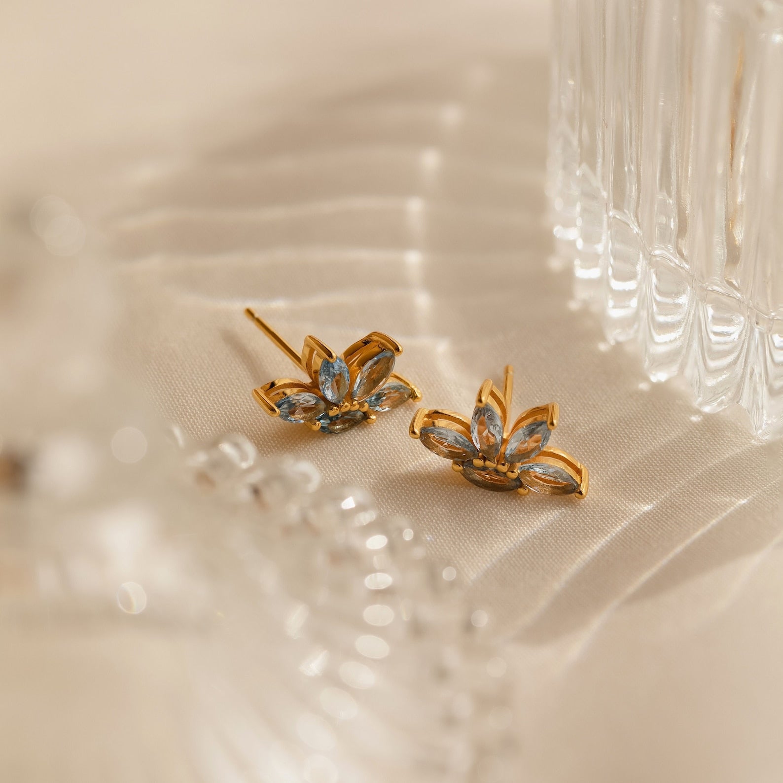 Aquamarine Marquise Studs with gold and blue floral design rest on cream fabric beside a clear glass object—ideal as a bridesmaids gift or for adding elegance to any special occasion.