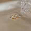 Marquise Diamond Drop Studs—gold earrings with clear, leaf-shaped gemstones inspired by Marquise-Cut Diamonds—rest on soft beige fabric beside a glass object.