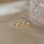 Marquise Diamond Drop Studs—gold earrings with clear, leaf-shaped gemstones inspired by Marquise-Cut Diamonds—rest on soft beige fabric beside a glass object.