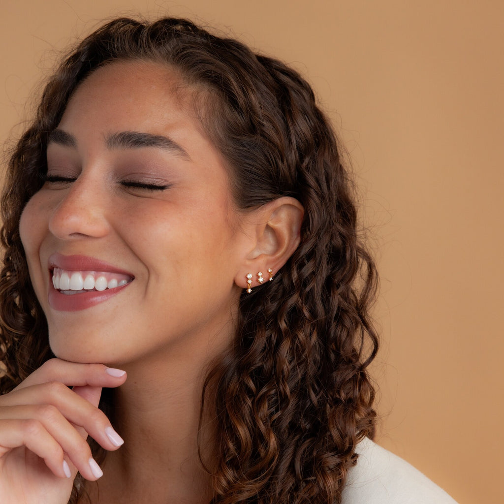 A smiling woman with curly hair and eyes closed gently touches her chin, showcasing the Triple Diamond Studs Set—elegant stacking earrings featuring three sparkling diamonds and delicate gold details.