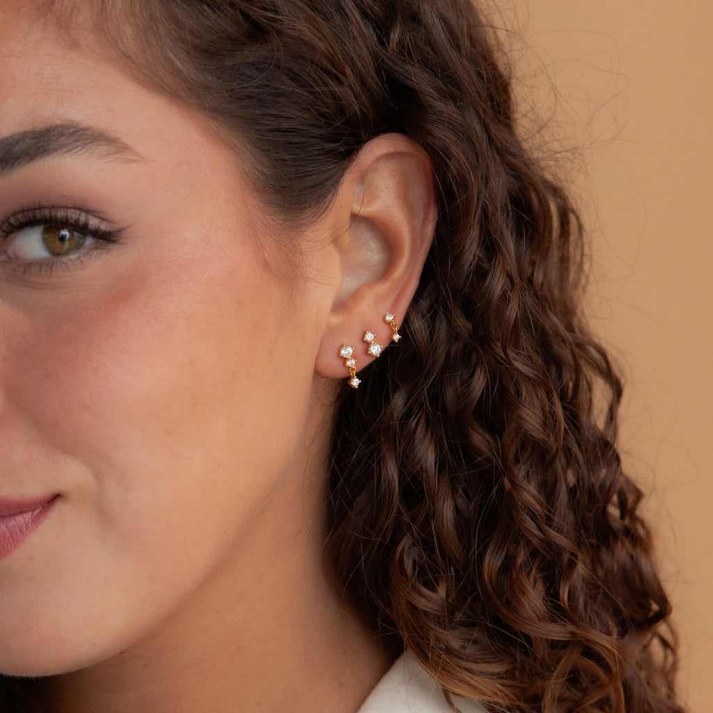 Woman with curly brown hair wearing the Triple Diamond Studs Set, along with gold and pearl stacking earrings.