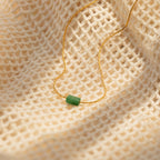 The Jade Bar Jewelry Set in 18K Gold features a gold necklace with a small green pendant, ideal for pairing with jade stud earrings, displayed on cream-colored textured fabric.