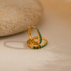 The Vera Pave Emerald Huggies, featuring green gemstones set in gold, rest on a light textured surface beside a smooth stone, adding vibrant color to these elegant earrings.