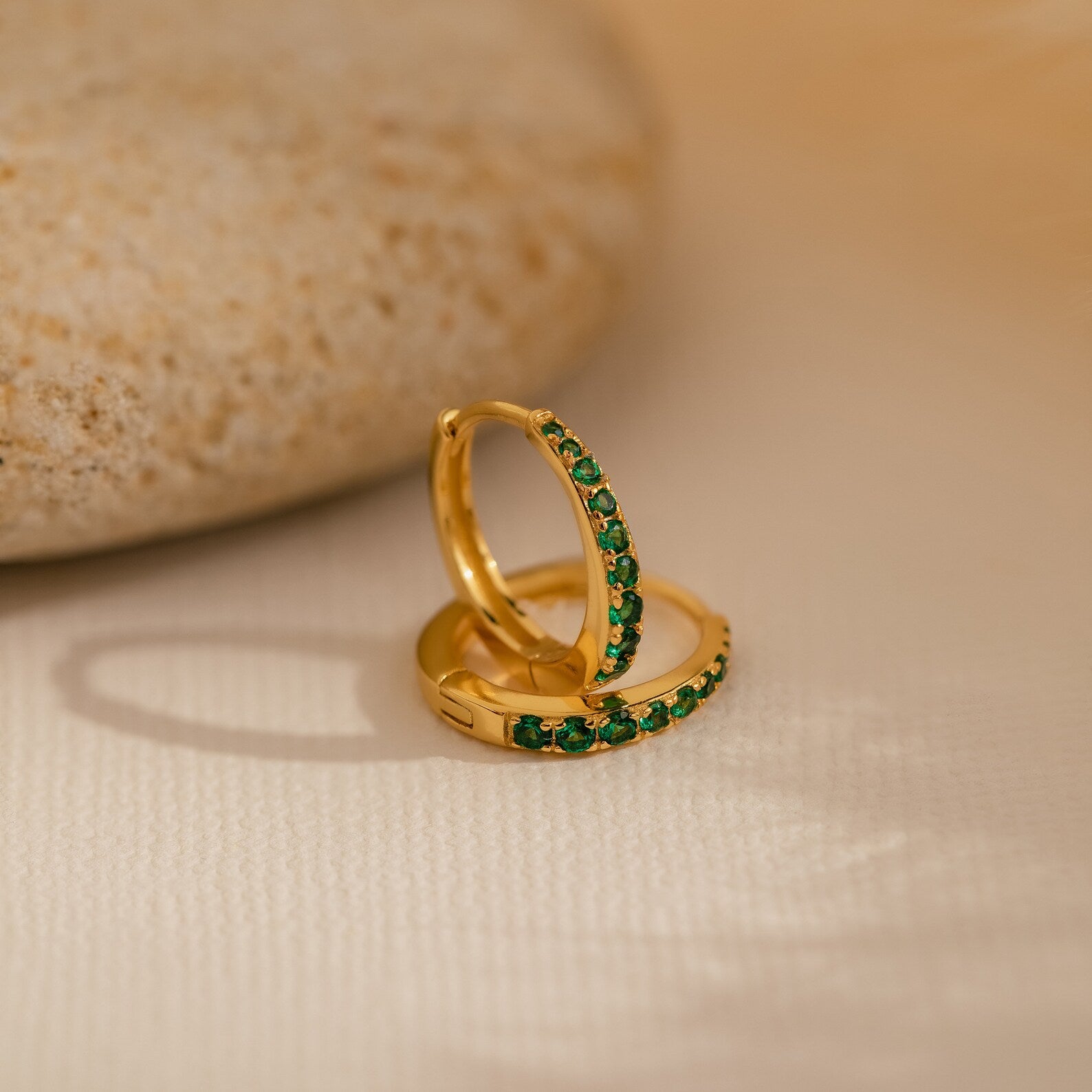 The Vera Pave Emerald Huggies, featuring green gemstones set in gold, rest on a light textured surface beside a smooth stone, adding vibrant color to these elegant earrings.