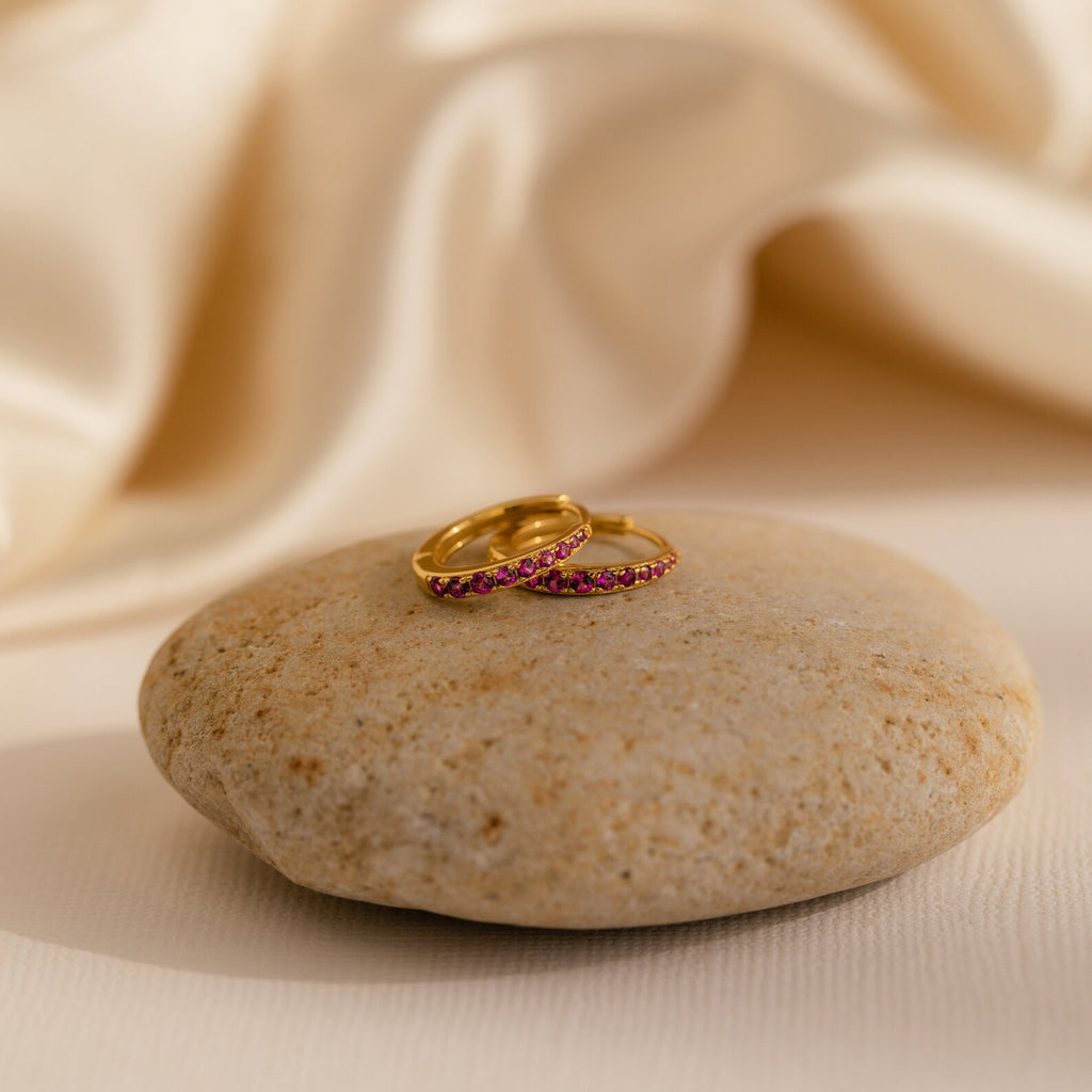 Two gold rings with pink stones rest on a smooth stone, set against beige fabric—pair them with the Vera Pave Ruby Huggies for an elegant, colorful accent.