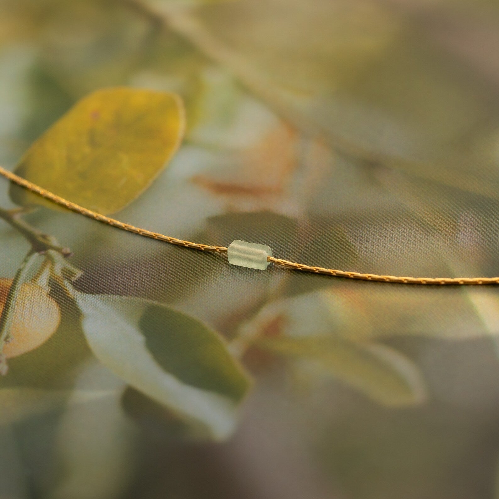 The Dainty Aventurine Bar Necklace features a thin gold cord and a small translucent aventurine bead, shown against a softly blurred natural background.