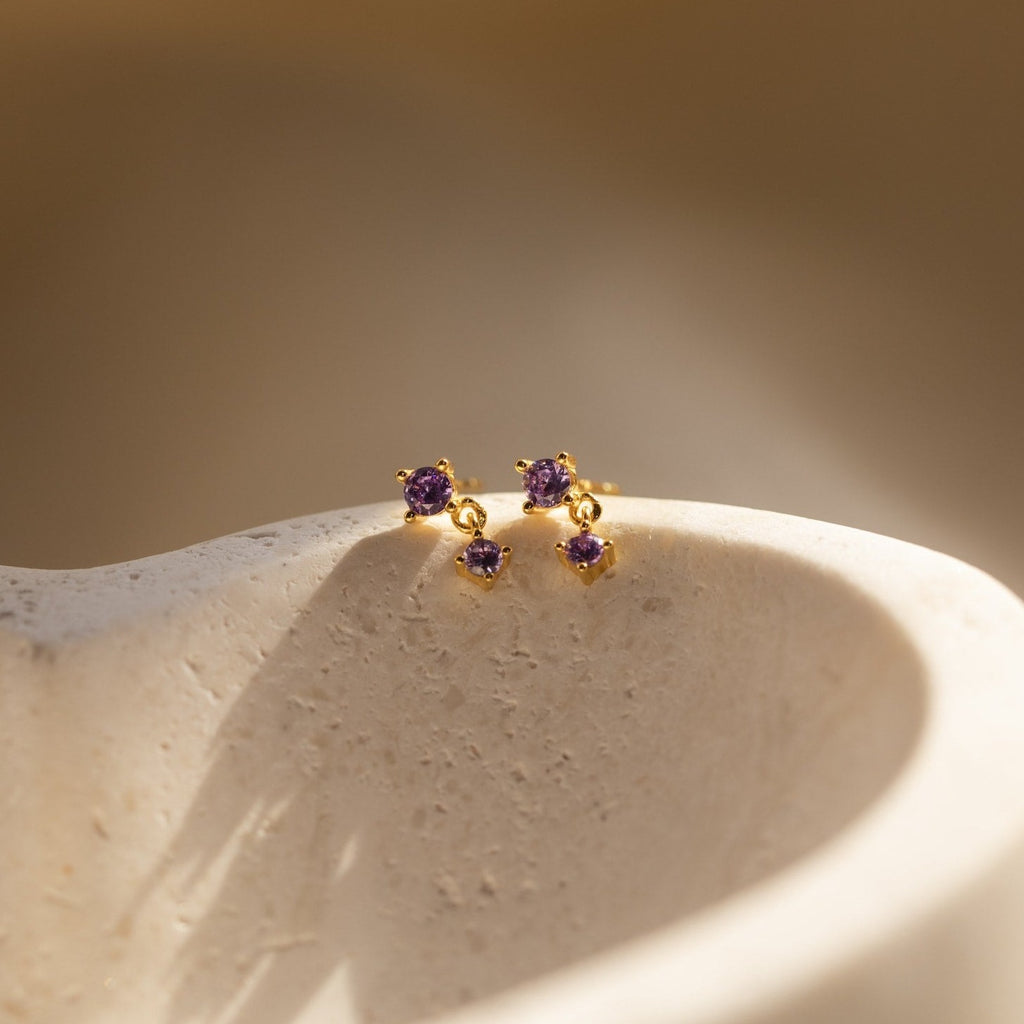 Amethyst Drop Studs with small purple gemstones rest on a textured, light-colored surface in soft sunlight.