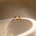 Amethyst Drop Studs with small purple gemstones rest on a textured, light-colored surface in soft sunlight.