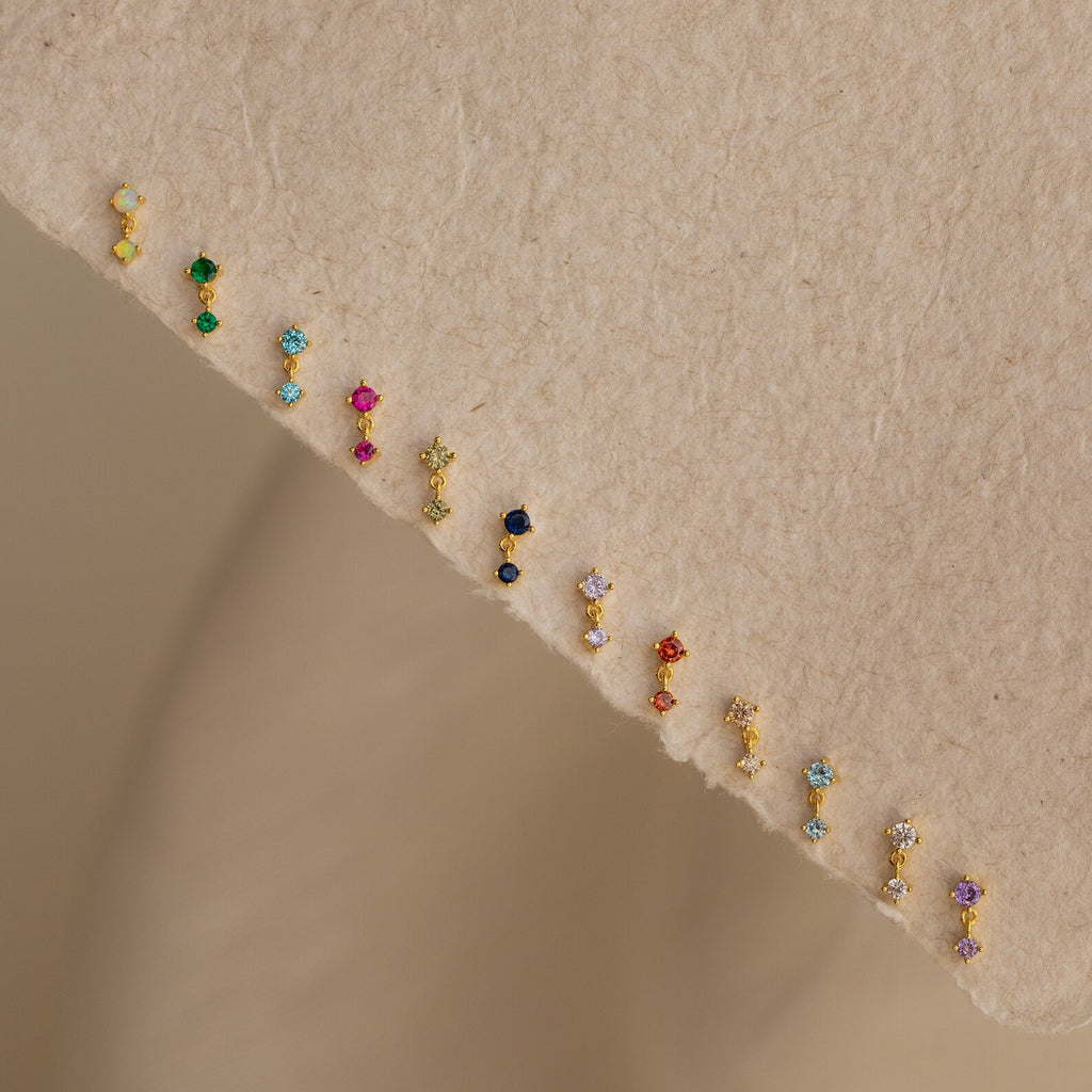 A pair of Amethyst Drop Studs is displayed on a beige textured surface, showcasing vibrant gemstone elegance perfect for gifting or everyday wear.