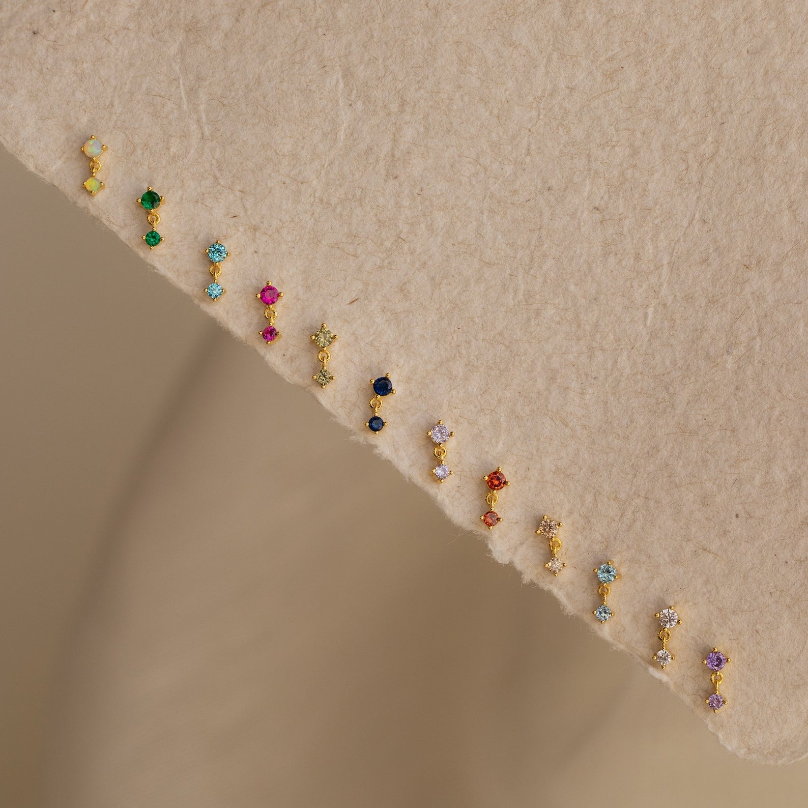 Emerald Drop Studs featuring vibrant gemstones set in a diagonal line, displayed on a beige textured surface.