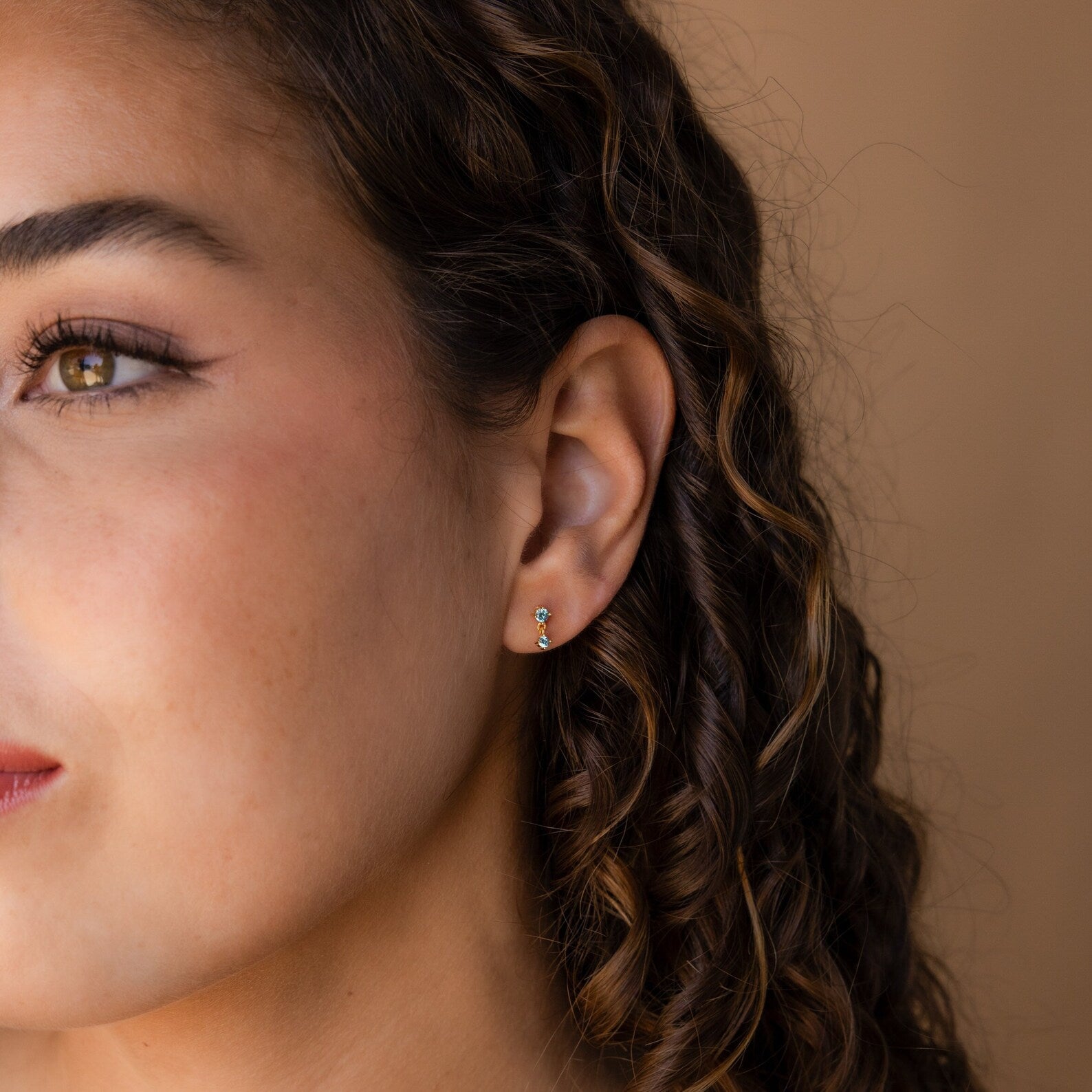 Woman with curly brown hair wears vibrant Amethyst Drop Studs, facing left against a beige background.