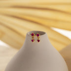 Ruby Drop Studs featuring small red gemstones are displayed on a white vase. The blurred beige background enhances these chic and elegant birthstone earrings, making them a perfect accessory.