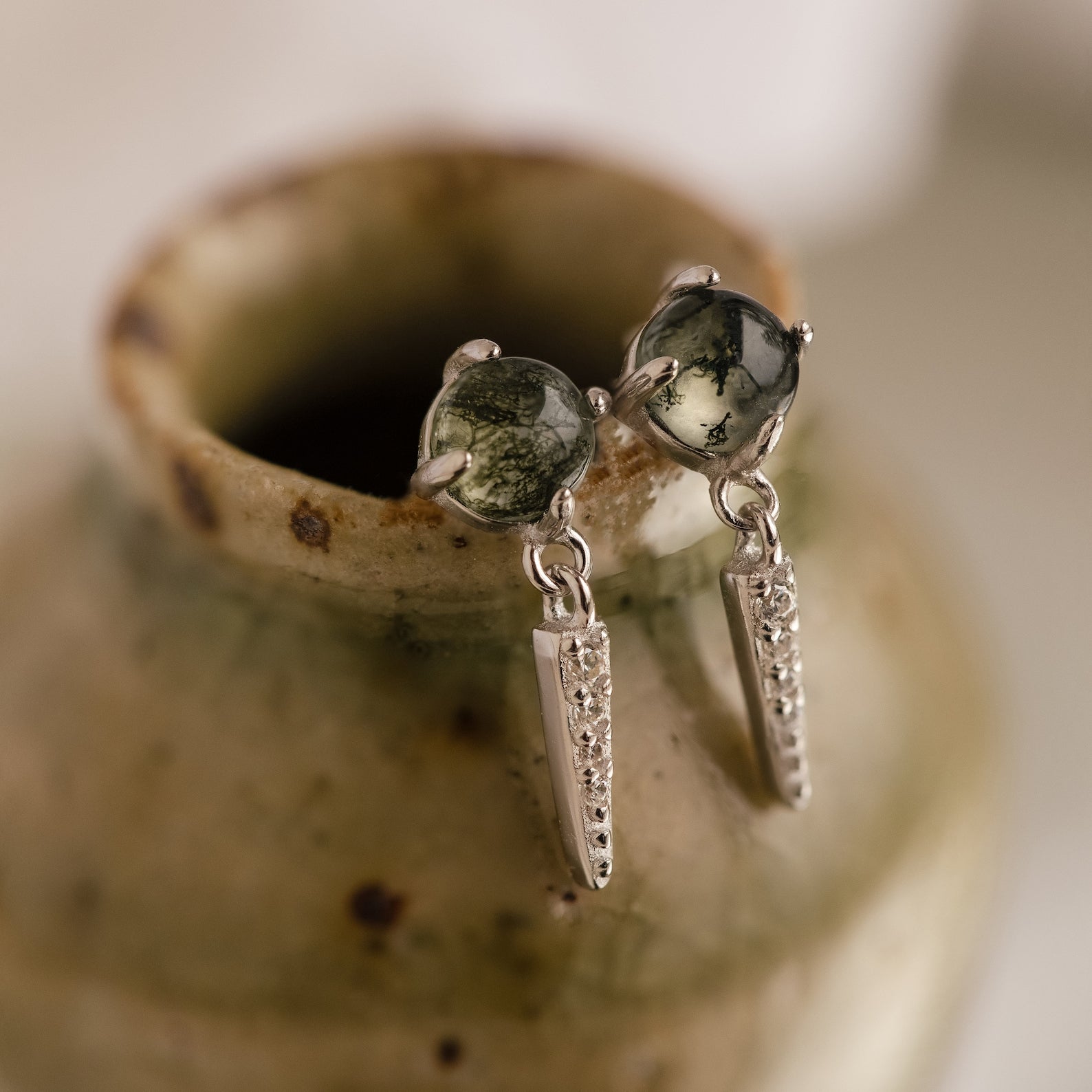 The Agate Diamond Drop Earrings feature silver dagger shapes with round moss agate stones and textured dangles, elegantly displayed on a ceramic vase.