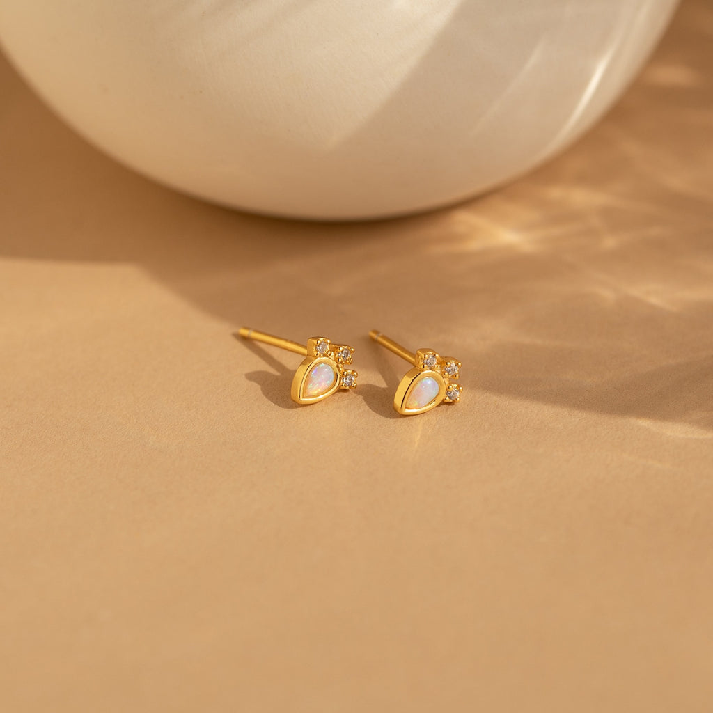 Dainty Opal Teardrop Studs—gold earrings featuring pearl teardrop centers and small crystals—are displayed on a beige surface, offering an elegant look.