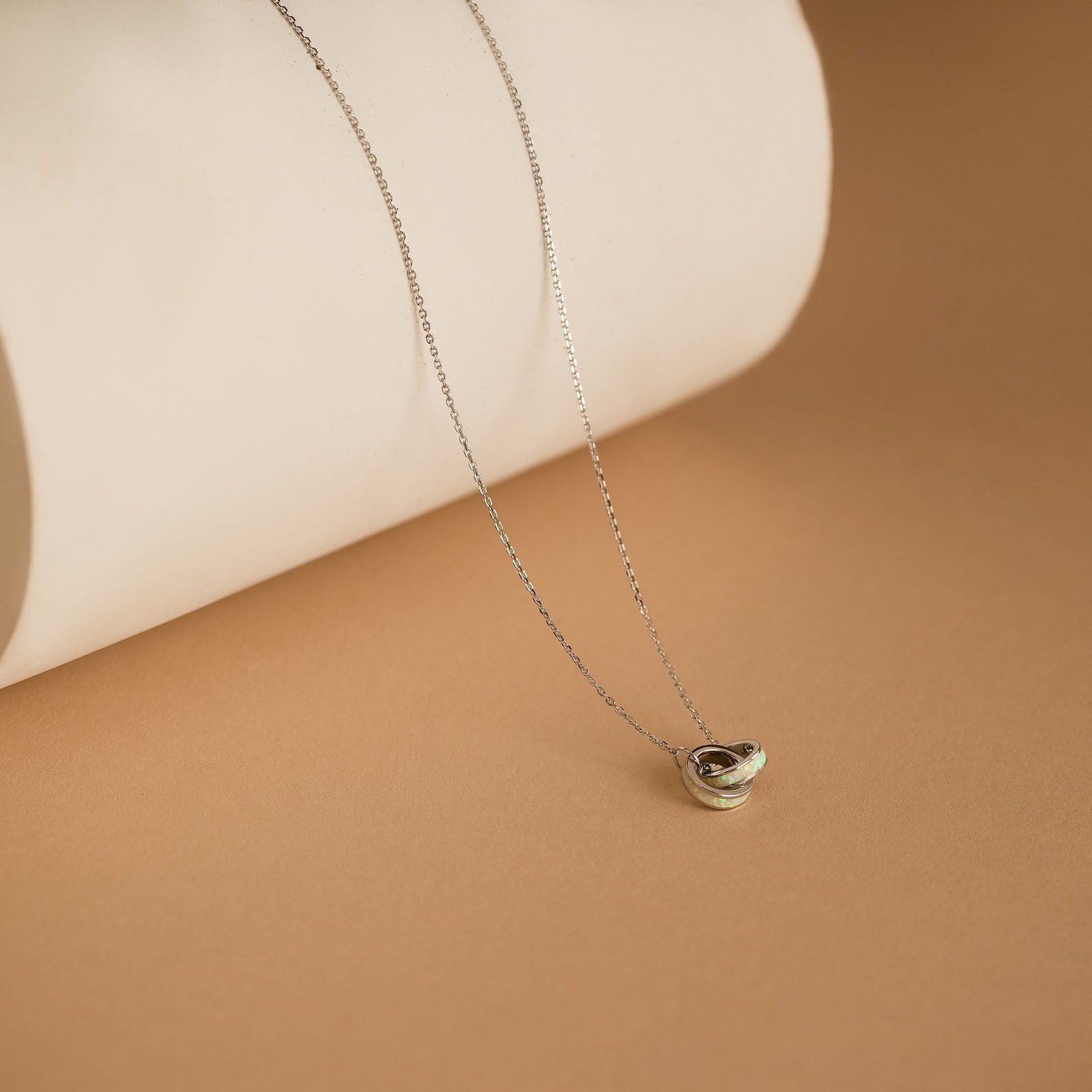 Interlocking Opal Inlay Necklace with a small pendant, showcased on a beige surface against a rolled white paper background—a stunning symbol of endless love, ideal for celebrating anniversaries.