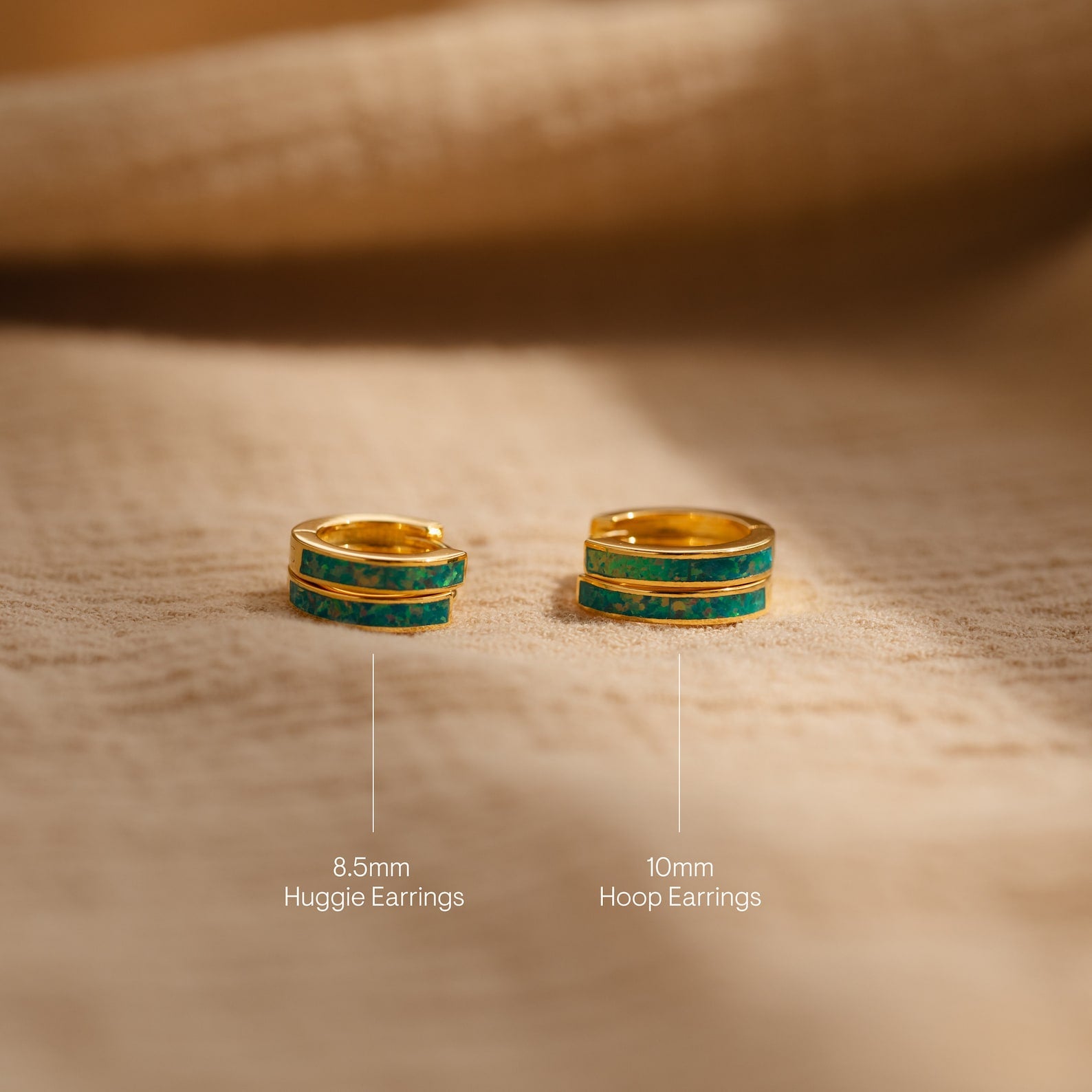 Two gold and turquoise earrings, including the Green Opal Inlay Hoops in 8.5mm huggie and 10mm hoop sizes, are displayed on a beige fabric surface.