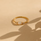 The Marquise Opal Diamond Ring features a gold band with a marquise opal center stone and two small diamonds on each side, displayed on a beige textured surface.