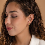 Woman with curly brown hair wearing Vera Pave Ruby Huggies, looking down with a soft smile against a beige background.