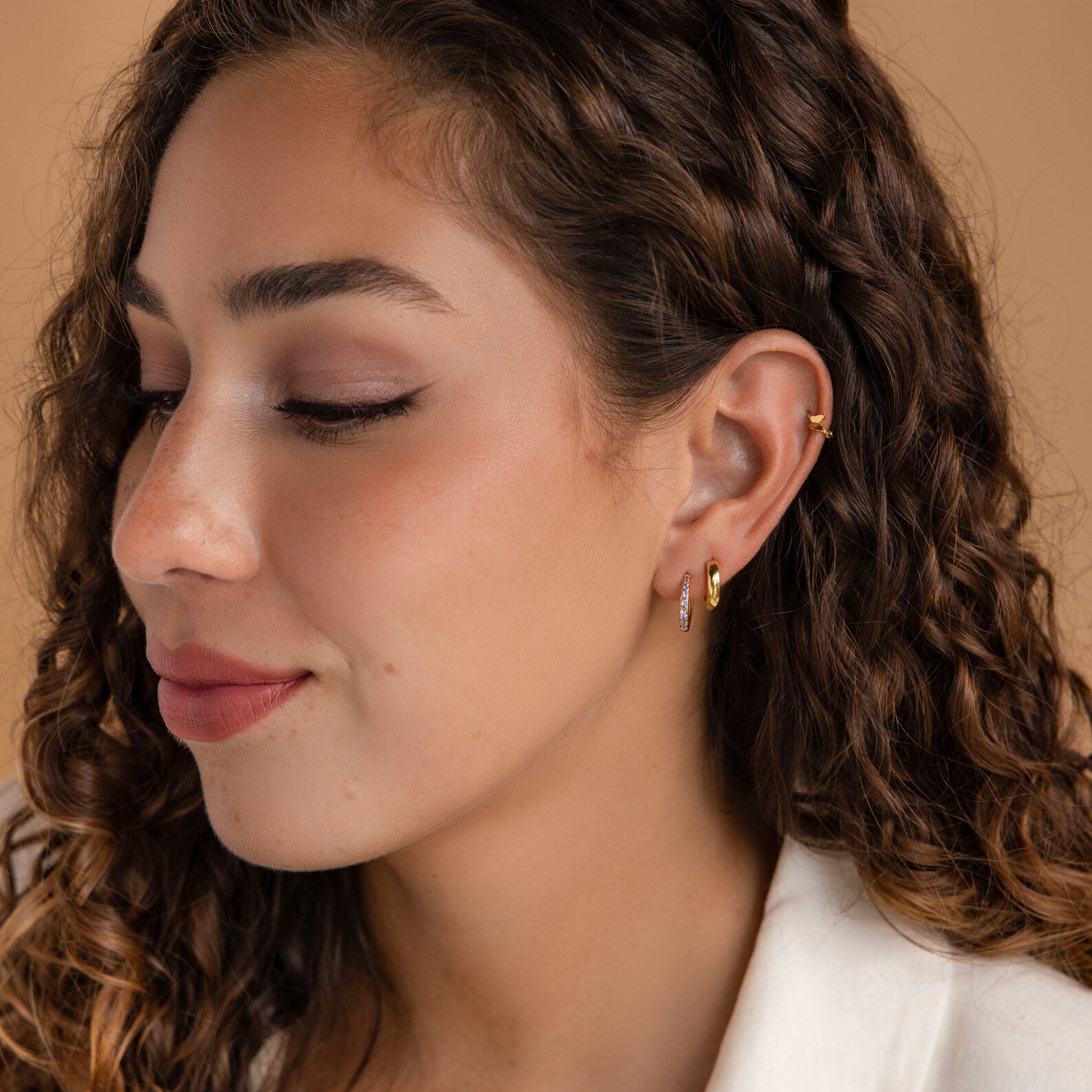 Woman with curly brown hair wearing Vera Pave Ruby Huggies, looking down with a soft smile against a beige background.