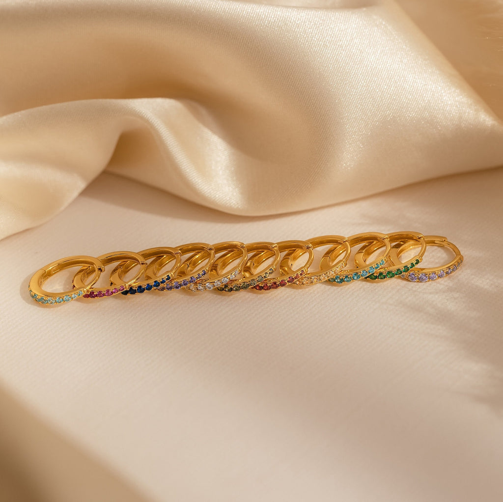 A row of gold rings, including Vera Pave Ruby Huggies with vibrant gemstones, displayed on beige satin fabric.